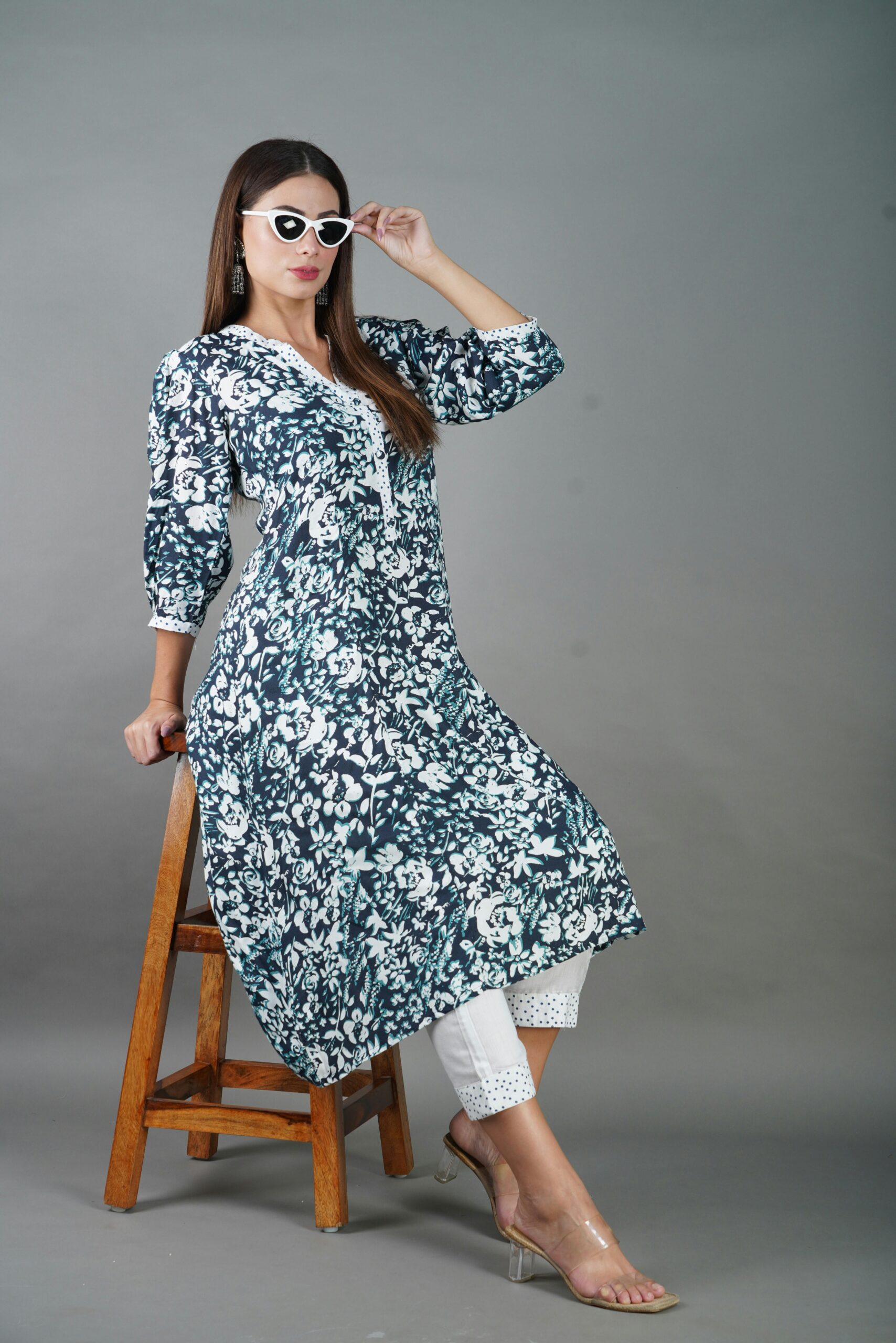 Fashionable woman wearing sunglasses and patterned dress posing elegantly on a stool.
