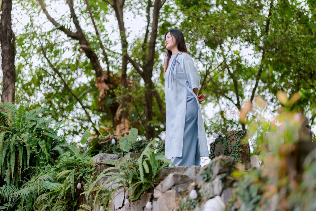 Woman in a blue dress standing peacefully amidst lush green foliage outdoors.